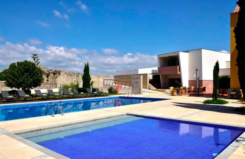 Em dia de sol, espreguiçadeiras, árvores, mesas, cadeiras e piscina em área de lazer externa de um dos hotéis no Algarve