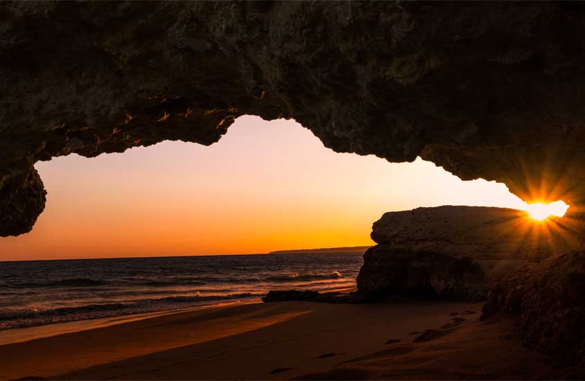 Pôr do sol visto de praia com paredões de pedra