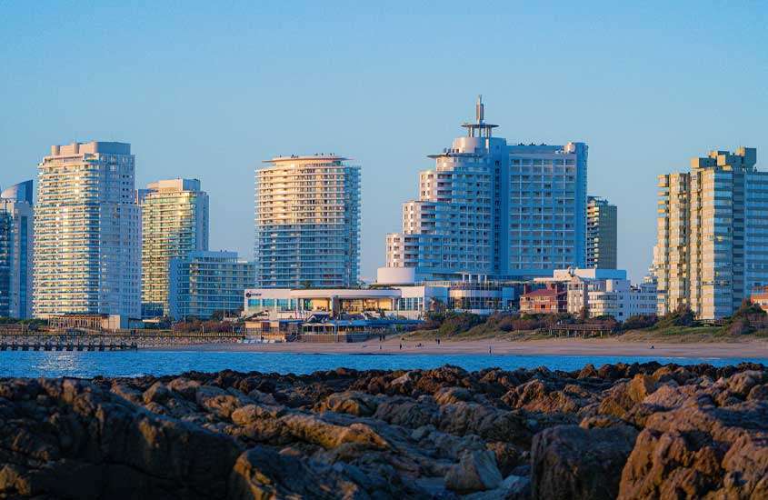 Em final de tarde, vista panorâmica de pedras e mar em praia. Ao fundo, prédios da cidade