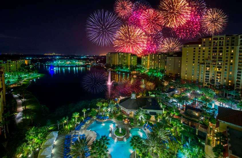 Durante a noite, vista aérea de área externa de hotel em Orlando com piscina, árvores ao redor. Ao fundo, fogos de artifício colorindo o céu