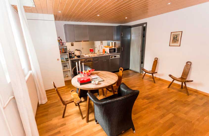 Sala e da cozinha conjugadas de apartamento em Berna, equipadas com mesa, cadeiras, eletrodomésticos, quadro, livros, armário e janela com cortina