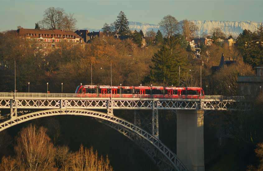 Durante dia de sol, trem passando em ponte. Ao fundo, árvores, prédios e montanhas