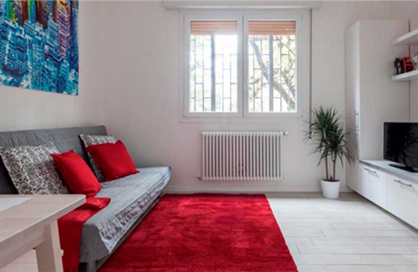 Em dia de sol, sala de estar com detalhes vermelhos, sofá, quadro, televisão, aquecedor e janela