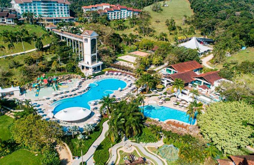 Durante dia de sol, vista aérea de um dos melhores hotéis fazenda do brasil com piscinas, guarda-sóis, espreguiçadeiras, árvores, áreas gramadas e prédios em volta