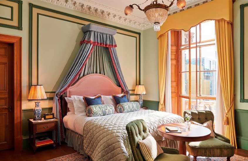 Quarto de hotel em Edimburgo com cama de casal, poltronas, mesa, luminárias, lustre, janela grande acortinada e tapete