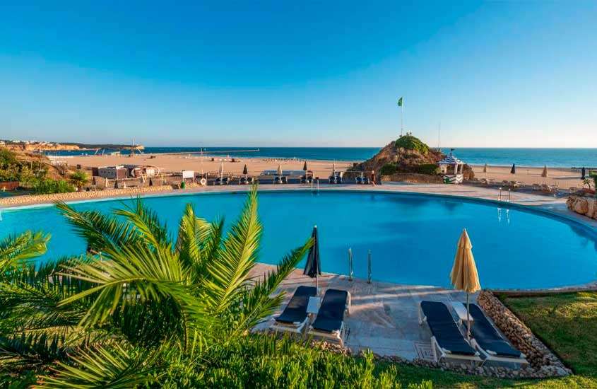 Em final de tarde, árvoeres e espreguiçadeiras em frente a grande piscina em área de lazer de hotel no Algarve com vista para o mar