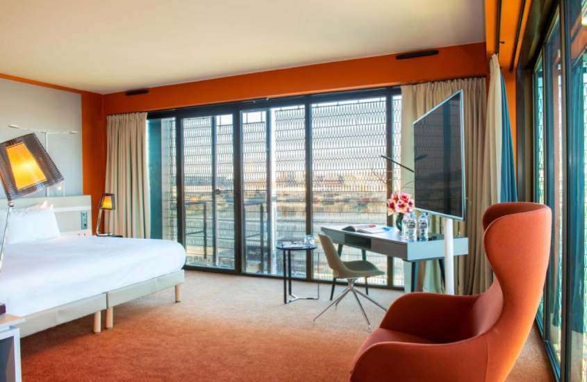 Quarto de hotel onde ficar em Bordeaux com cama de casal, cadeiras, área de trabalho TV, luminárias e varanda com cortina