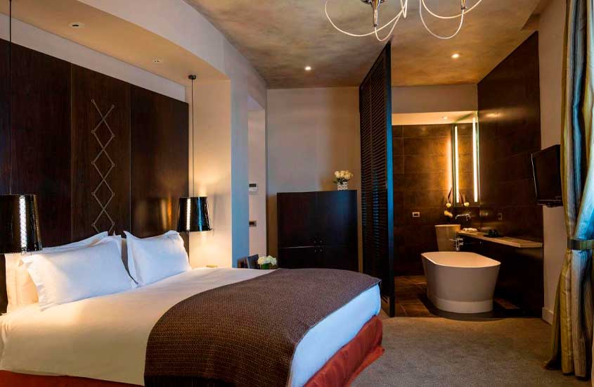 Quarto de hotel onde ficar em Montevideu com cama de casal, luminárias, armário, banheiro separado por estrutura de madeira, banheira, espelho, televisão e flores