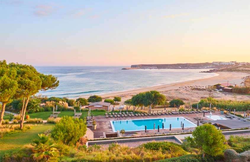 Em final de tarde, vista aérea de árvores e espreguiçadeiras ao redor de piscina em área de lazer externa de hotel no Algarve com vista para o mar