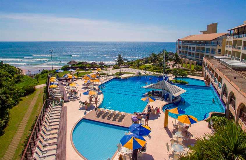 Durante o dia, vista aérea de guarda-sóis e espreguiçadeiras ao redor de piscina em área de lazer de resort com vista para o mar