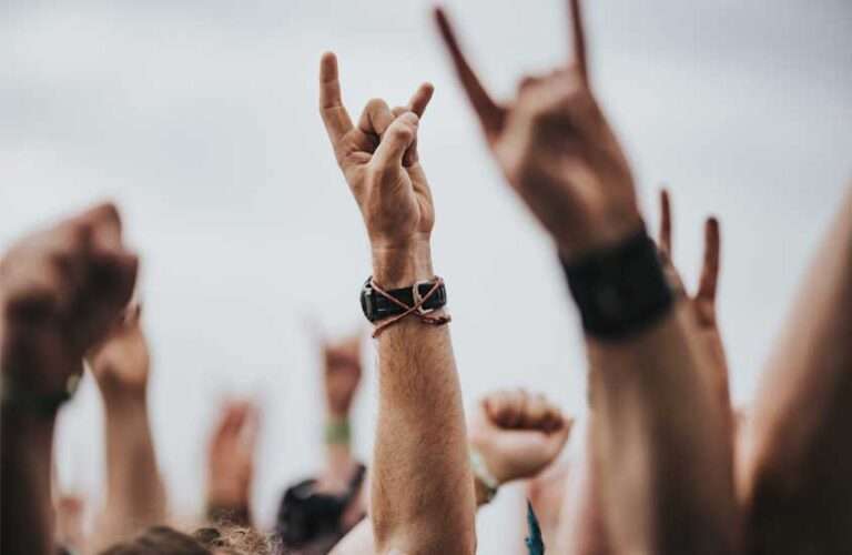 Em dia nublado, braços levantados com pulseiras e relógio, fazem símbolo de rock