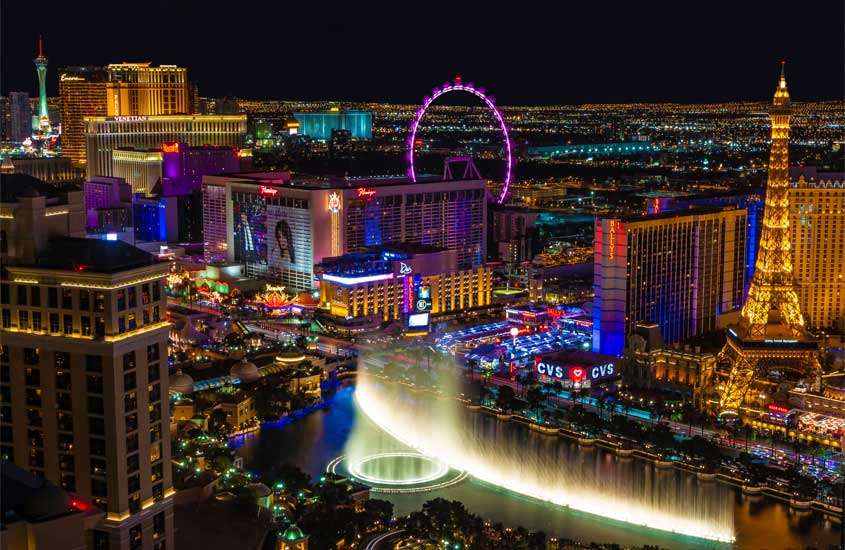 Durante a noite vista aérea panorâmica de Las Vegas, cenário de filme Se beber não case, com vários prédios iluminados, roda gigante, lago artificial, réplica de torre eiffel e carros na rua