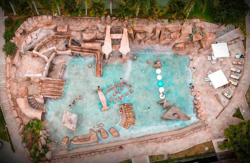 Durante o dia vista aérea de uma piscina com decorações em pedra, rodeada de espreguiçadeiras e guarda-sóis
