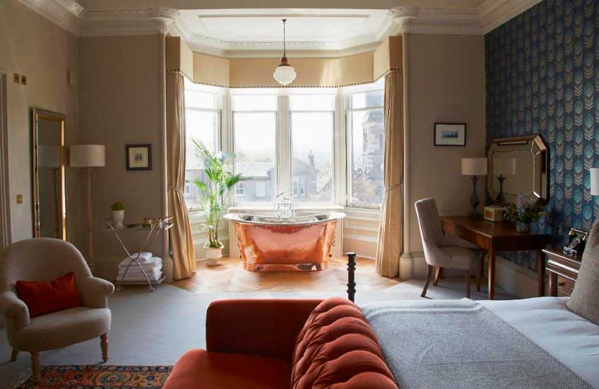 Durante o dia, quarto de um hotel em Edimburgo com cama de casal, poltrona, banheira de cobre, espelhos, sofá, luminárias, janelas grandes, suporte de toalhas e vasos de planta