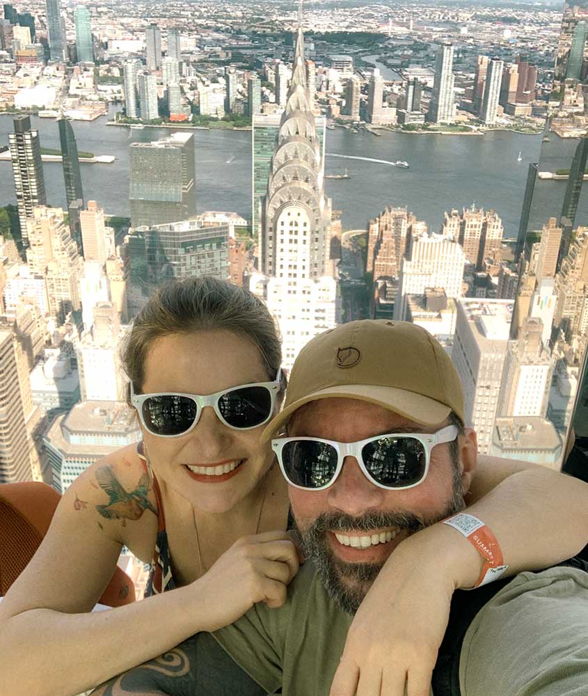 Durante o dia, Bárbara Rocha e Vagner Alcantelado, ambos usando óculos de sol, sorriem abraçados para a foto. Ao fundo, vista panorâmica de Nova York