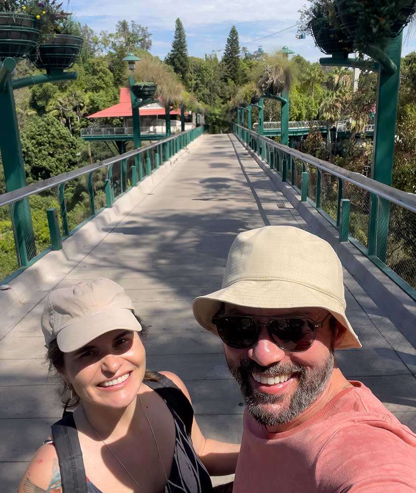 Em um dia ensolarado, Bárbara Rocha, com boné, cabelo preso e regata, e Vagner Alcantelado, com chapéu, óculos de sol e blusa, sorriem para uma selfie no Zoológico de San Diego.