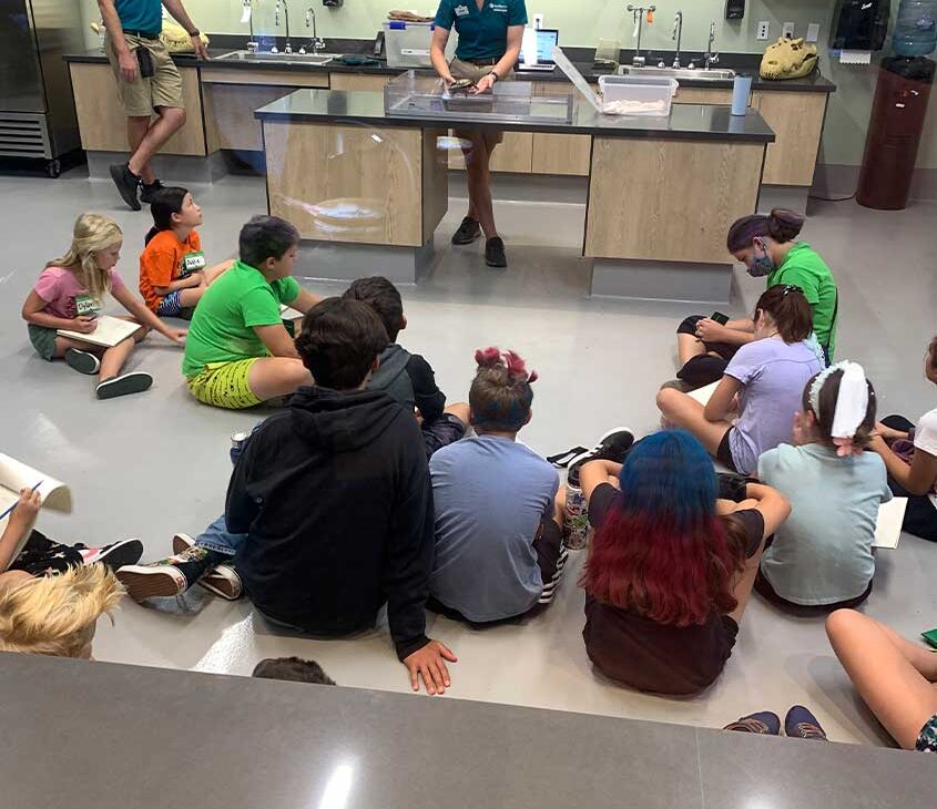 Diversas crianças sentadas no chão de um laboratório no Zoológico de San Diego, atentamente observando os funcionários em uma oficina educativa interativa
