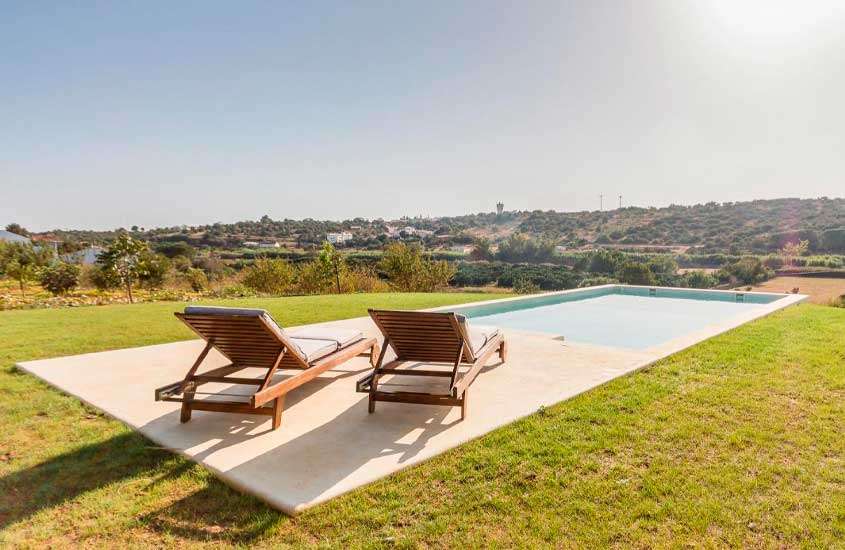 Em dia de sol, espreguiçadeiras de madeira em frente a piscina em quintal de uma das casas de férias no Algarve com vista para as montanhas