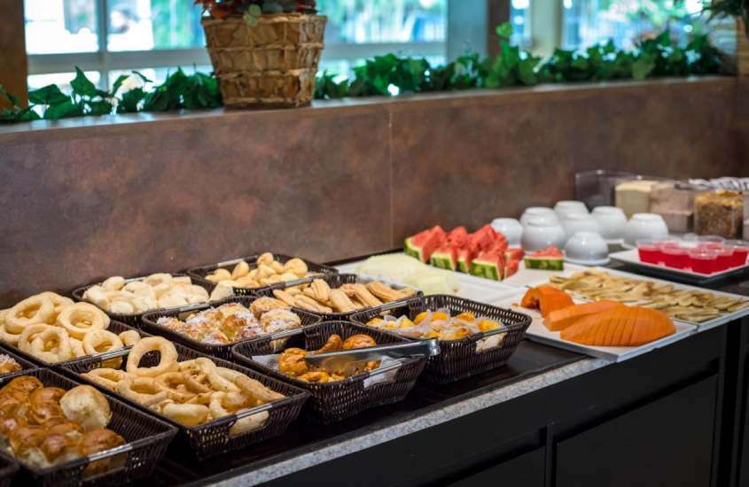 Mesa de café da manhã de um hotel para lua de mel em caldas novas com biscoitos, pães, churros, frutas, xícaras, gelatina, granola, leite em pó, achocolatado e plantas ao redor