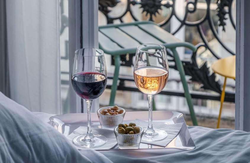 Taças de vinho acompanhadas de azeitonas e amendoins, em cima de cama de quarto de hotel onde ficar em Bordeaux com varanda e cadeiras