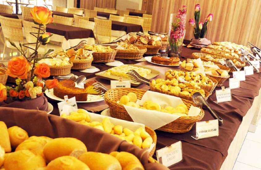 Mesa de café da manhã de um hotel para lua de mel em caldas novas com pães, salgados, bolos, broa e flores. Ao lado, mesas e cadeiras