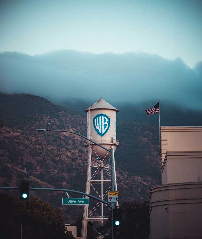 Em um dia nublado, destaque para um sinal de trânsito verde em primeiro plano, enquanto ao fundo há uma placa com o símbolo dos Estúdios Warner Bros. e a paisagem de montanhas.