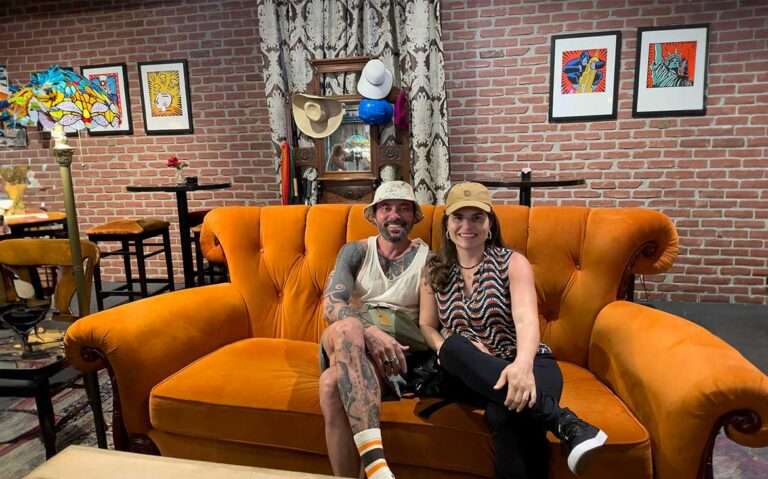 Vagner Alcantelado e Bárbara Rocha, sentados em um sofá, sorriem capturando um momento descontraído no ambiente do warner bros. studio tour hollywood