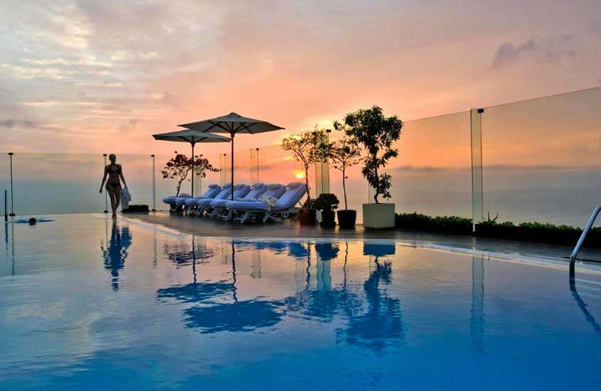 Em final de tarde, área de lazer com piscina, espreguiçadeiras, guarda-sóis, vasos de planta e vista do mar em um dos melhores hoteis em lima