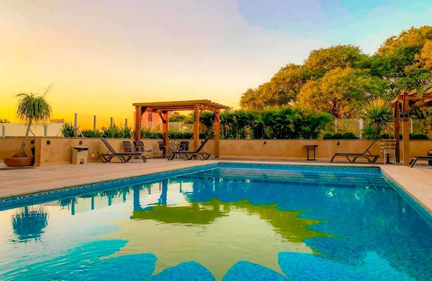 Em final de tarde, área de lazer de um hotel em foz do iguaçu com piscina, espreguiçadeiras, árvores e vegetação ornamental ao redor