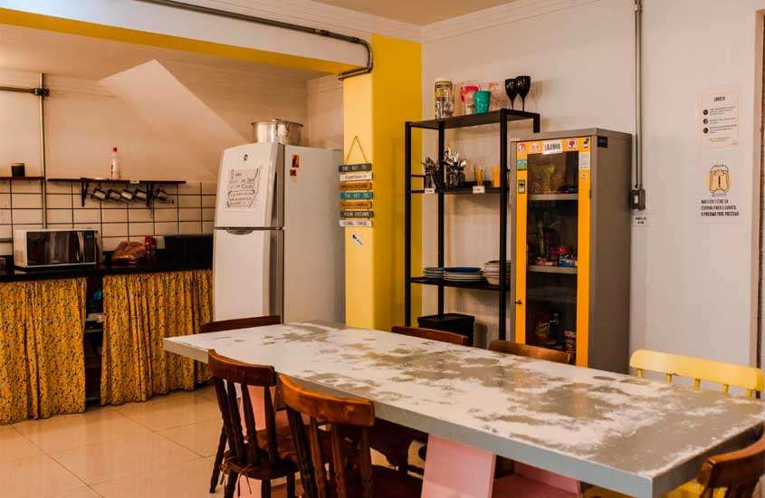 Sala de hostel com mesa, cadeiras, armários, pia de cozinha integrada e geladeira