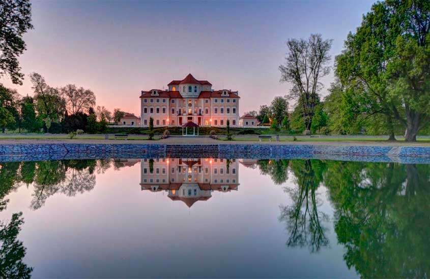 Durante o anoitecer, fachada de um dos castelos europeus com lago em frente refletindo a paisagem, árvores, bancos e parte gramada ao redor