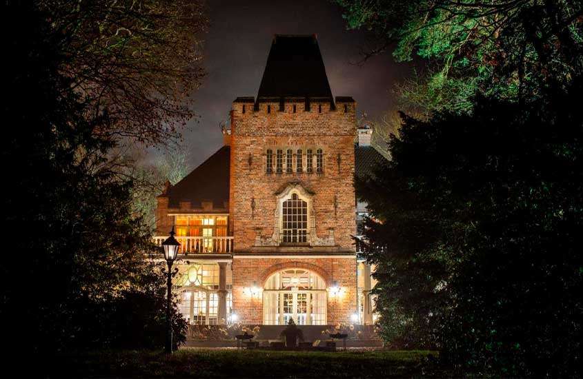 Durante a noite, fachada de um dos hotel em castelo iluminado e com árvores em volta