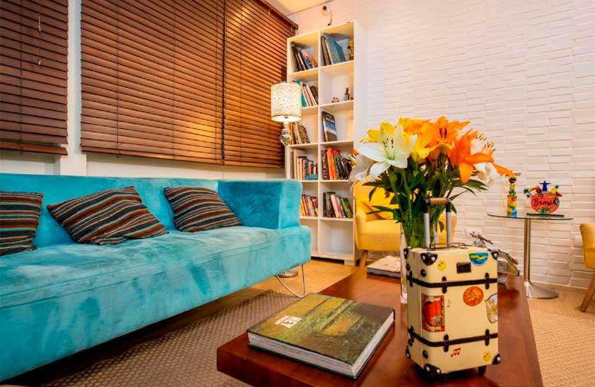 Lounge de um hotel pet friendly são paulo com sofá, mesas, prateleira de livros, poltronas, janela com persiana e flores decorativas
