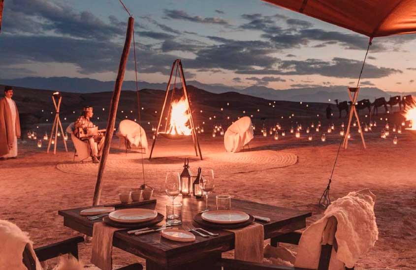 Durante a noite, jantar em deserto com velas, fogueira, mesa posta, camelos e cadeiras com pelego