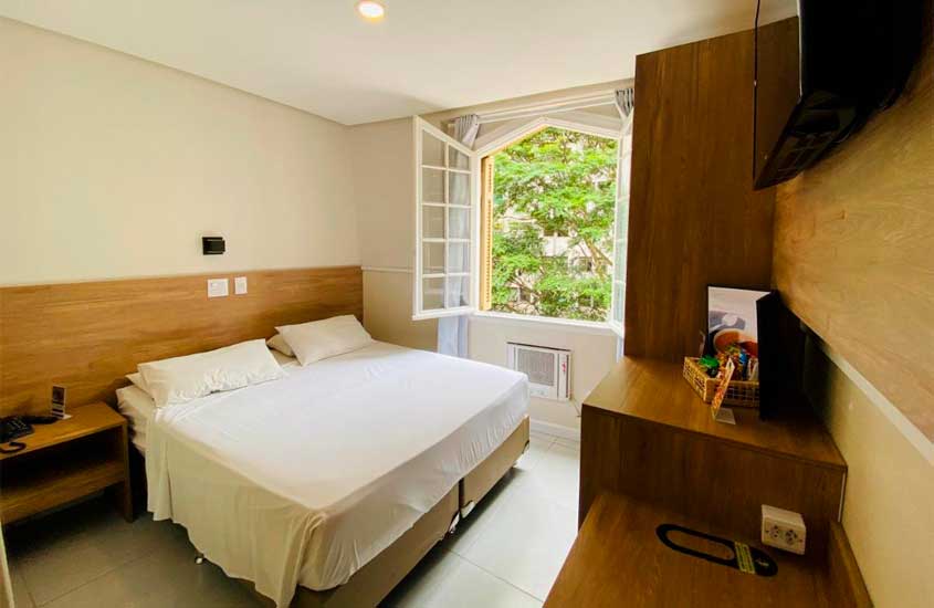 Durante o dia, quarto de hotel pet friendly são paulo com cama de casal, TV, armário, criado, ar-condicionado e janela acortinada com vista da árvore