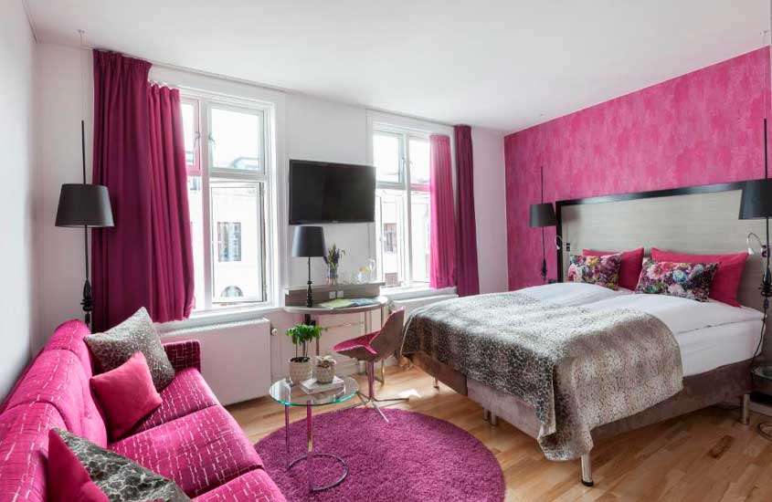 Quarto de hotel em copenhagen com decoração em tons de rosa, cama de casal, sofá, mesa, área de trabalho, TV, janelas acortinadas, tapete e luminárias