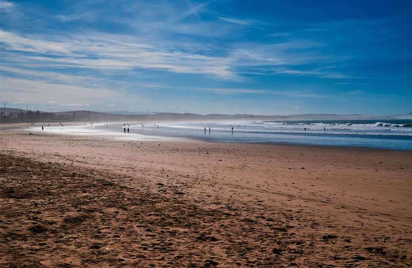 Em dia de sol, pessoas andando em areia de praia. Ao fundo, mar
