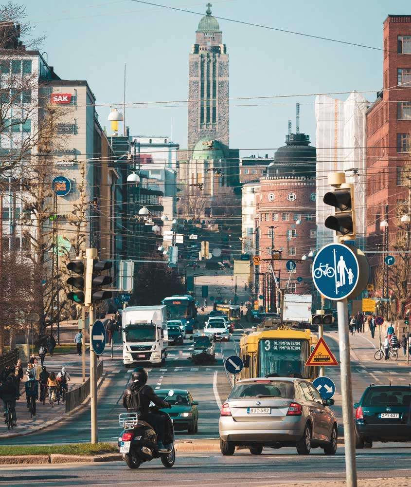 Em dia de sol, rua de um dos bairros onde ficar em helsinque com carros, motos, prédios e pessoas