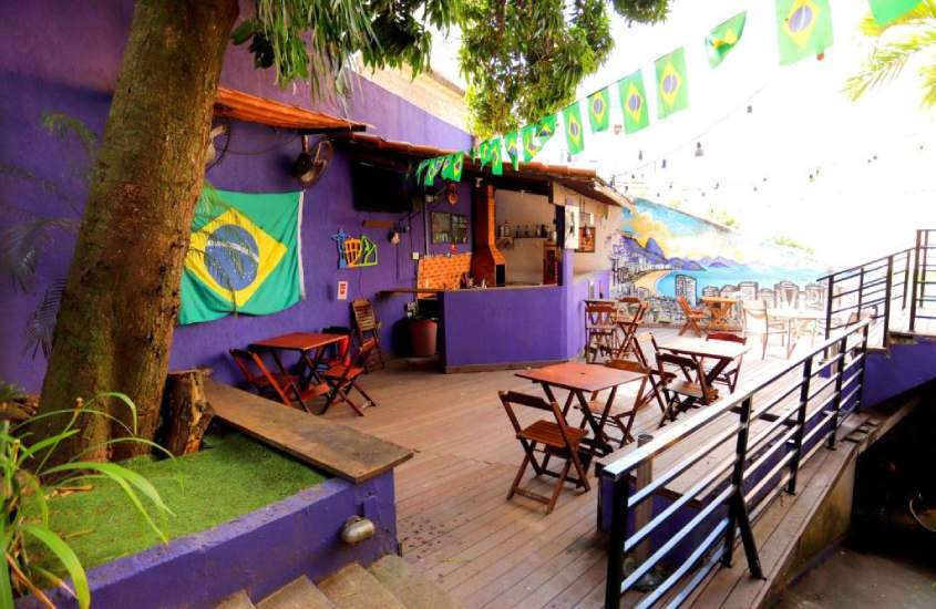 Em um dia de sol, área de lazer de hostel em copacabana barato com bancos, cadeiras, mesas, área de churrasqueira e decoração de bandeira do brasil