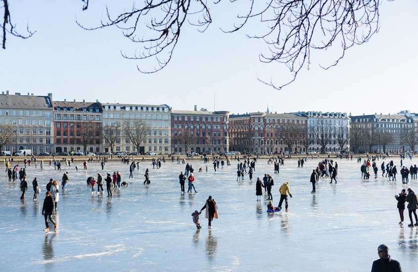 Em dia de sol, pessoas patinando em lago congelado, em Nørrebro, um bairro onde ficar em copenhagen. Ao fundo, árvores e prédios.