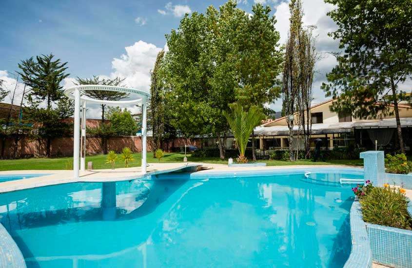 Em um dia de sol, área de lazer de um hotel em cusco com piscina, parte gramada, árvores, flores decorativas e hotel atrás