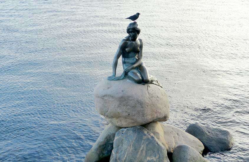 Em final de tarde, pássaro em cima de cabeça de escultura de sereia em cima de pedras em frente ao mar