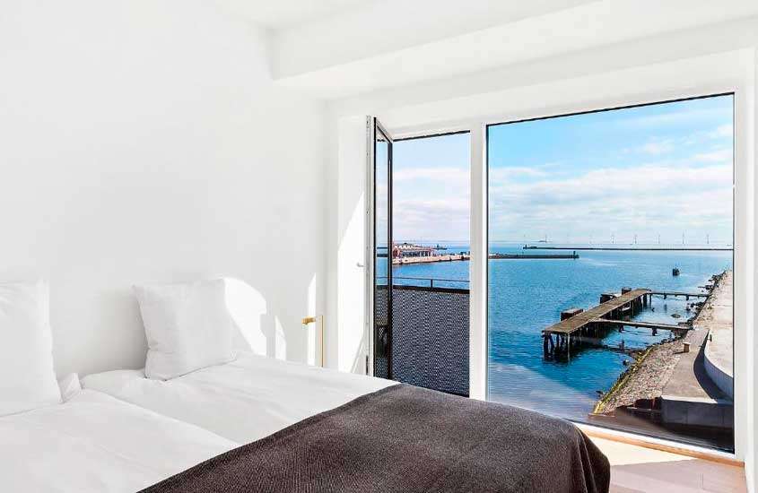 Em dia de sol, quarto de um hotel onde ficar em copenhagen com janela grande com vista para o mar, cama de casal e varanda
