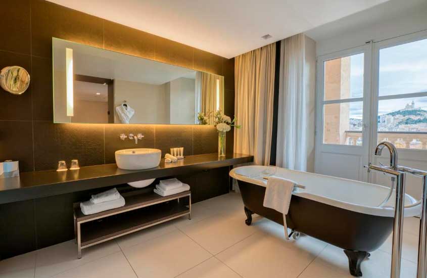 Banheiro de hotel com banheira, toalhas, espelhos, vaso decorativo e janela grande acortinada com vista para a cidade