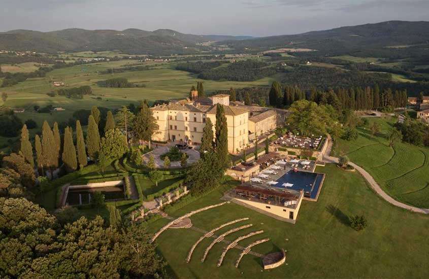 Em dia de sol, vista aérea de um dos castelos europeus com entorno gramado, árvores, piscina, carros, mesas cadeiras, guarda-sóis