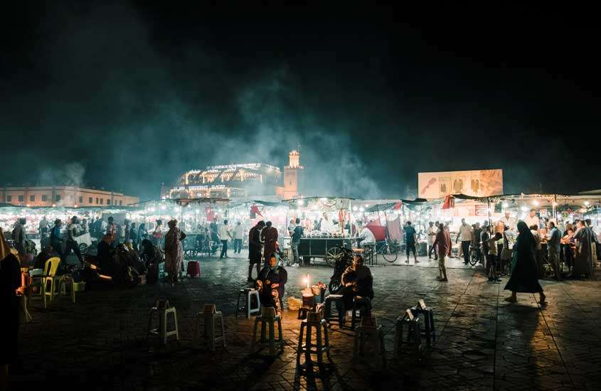 Durante a noite, mercado com várias pessoas, barracas e construções atrás