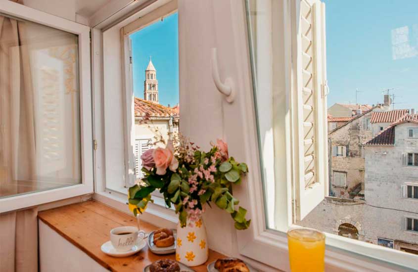 Parte da janela de um hotel onde se hospedar em split com vista da cidade, flores decorativas, cortina, suco de laranja, cafe, donut e pães