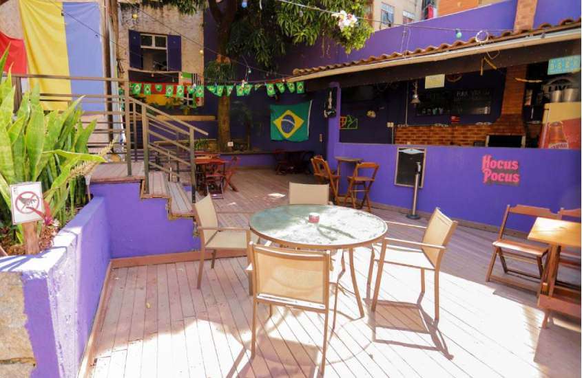 Em um dia de sol, área de lazer de um hostel em copacabana com mesas, cadeiras, flores decorativas e bandeira do brasil no fundo
