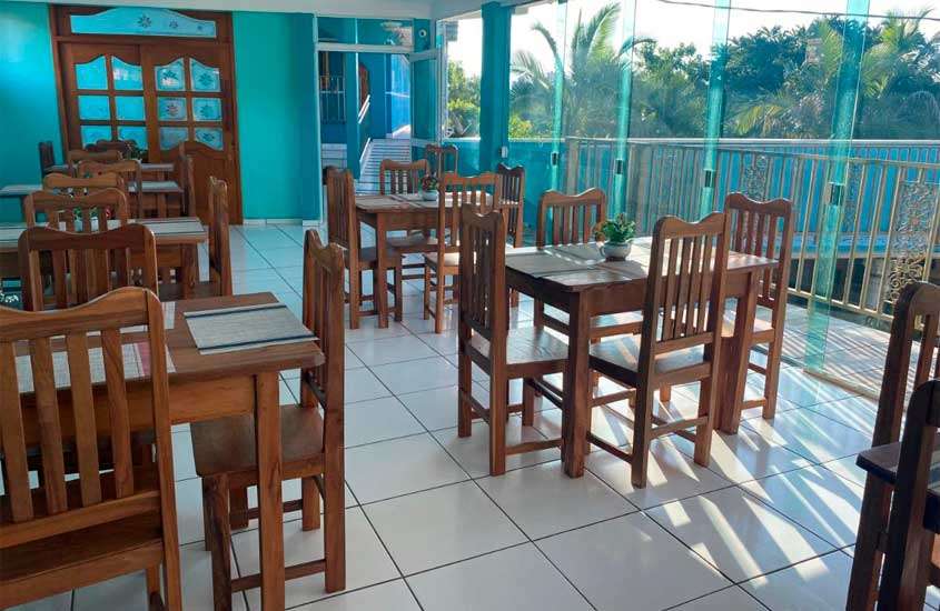 Em tarde de sol, área de café da manhã de hotel com mesas, cadeiras, plantas decorativas e árvores atrás