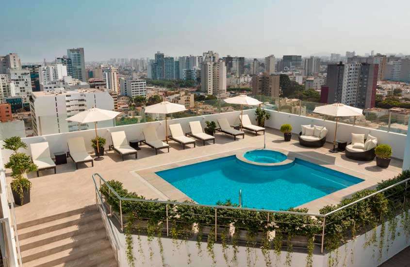 Em dia de sol, área de lazer de um dos melhores hotéis em Lima com piscina, jacuzzi, espreguiçadeiras, sofás, guarda-sóis, plantas e vista da cidade
